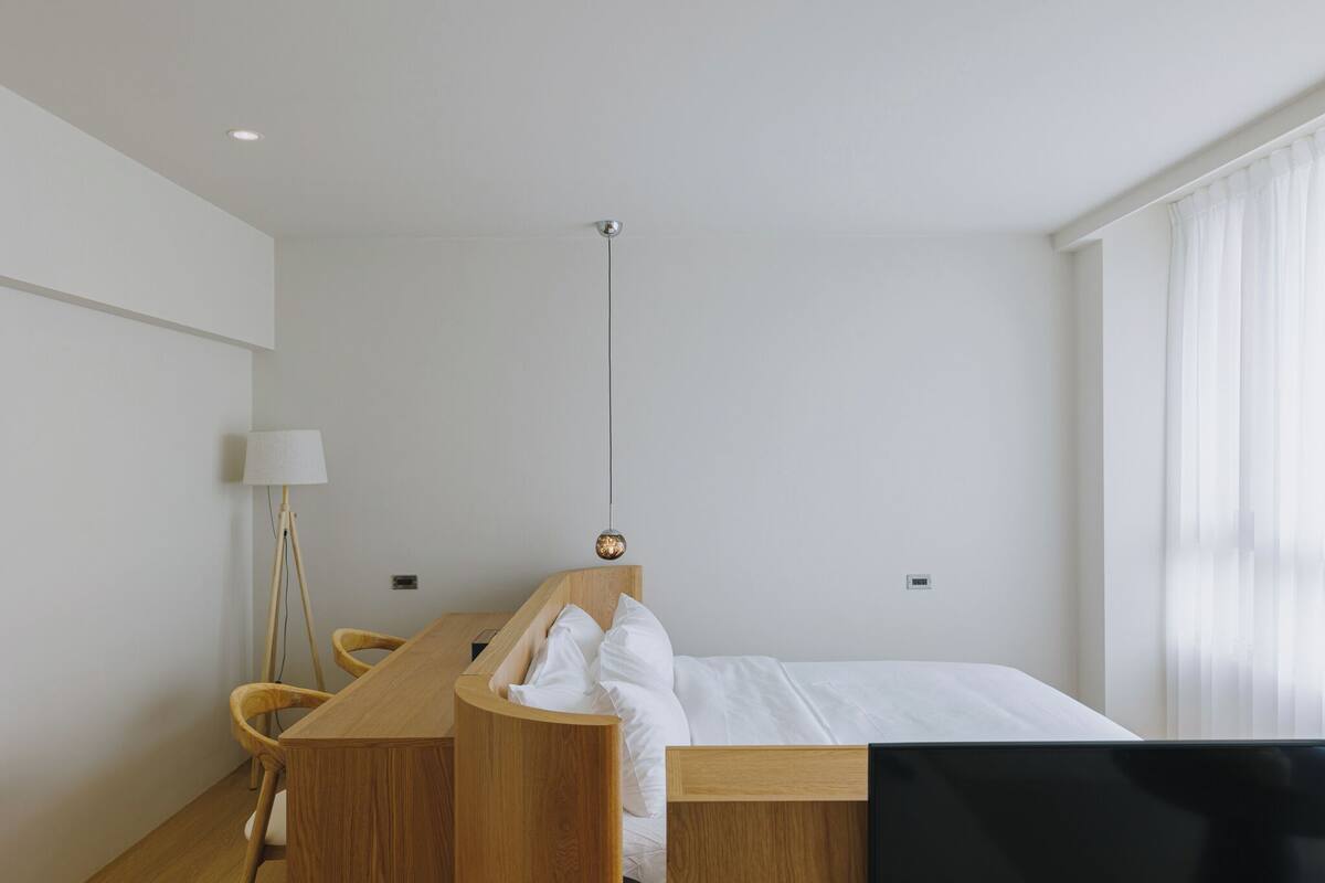 Habitación de hotel minimalista con escritorio de madera, una cama y una lámpara de pie.