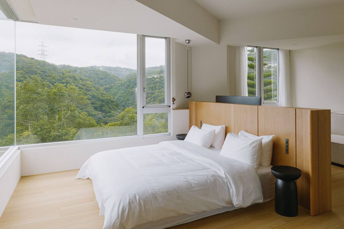 Un dormitorio con una cama grande, cabecera de madera y vistas a un paisaje boscoso a través de amplias ventanas.