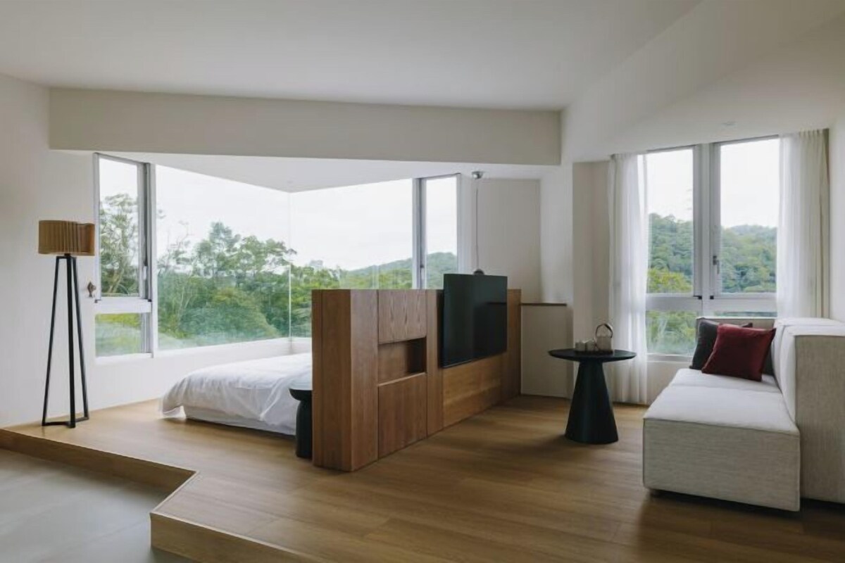A modern hotel room with a large bed, a wooden entertainment unit, a small side table, and a sofa.