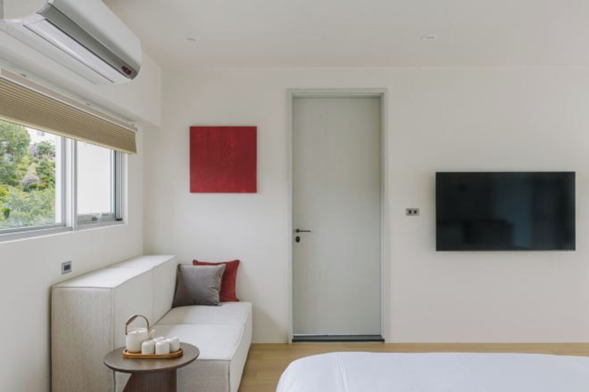 A modern hotel room with a bed, a sofa, a small table with a tray of candles, a TV mounted on the wall, and a door.