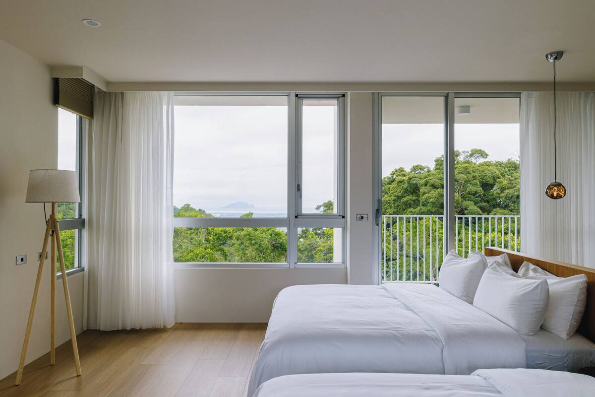 Un dormitorio con una cama grande, una lámpara de pie y vistas a zonas verdes a través de las ventanas.