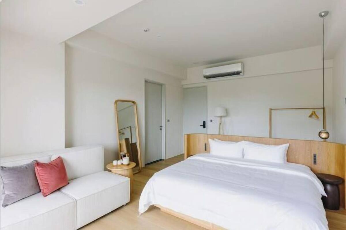 Habitación de hotel con cama, sofá blanco, espejo, mesita de noche y equipo de aire acondicionado.