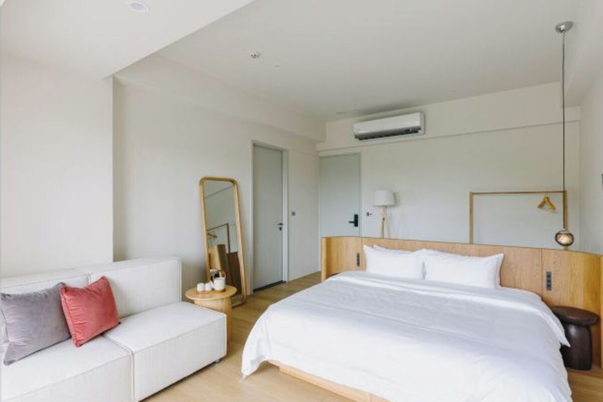 A hotel room with a bed, a white sofa, a mirror, a bedside table, and an air conditioning unit.