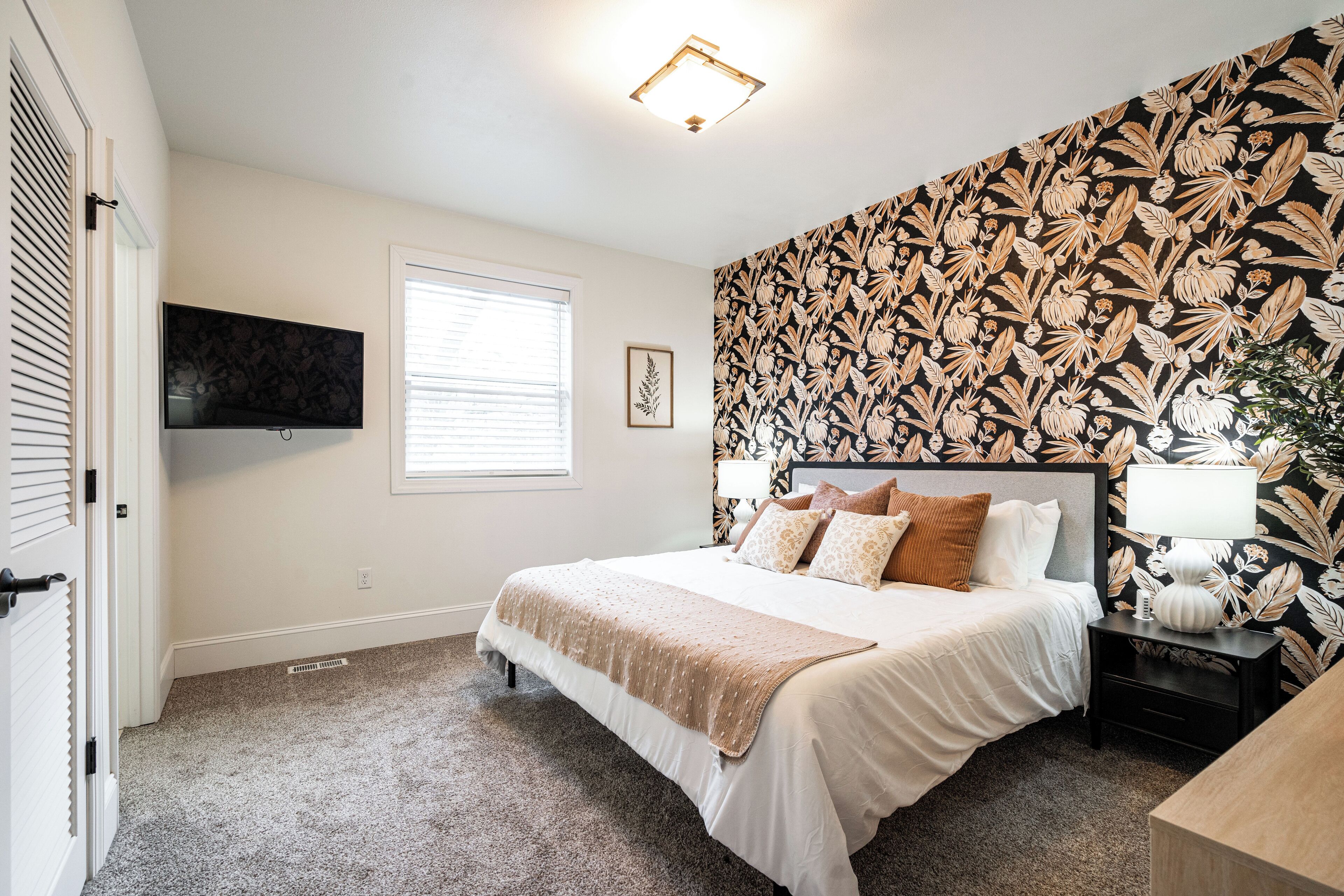 A bedroom with a bed, a TV mounted on the wall, a window, and a patterned wallpaper.