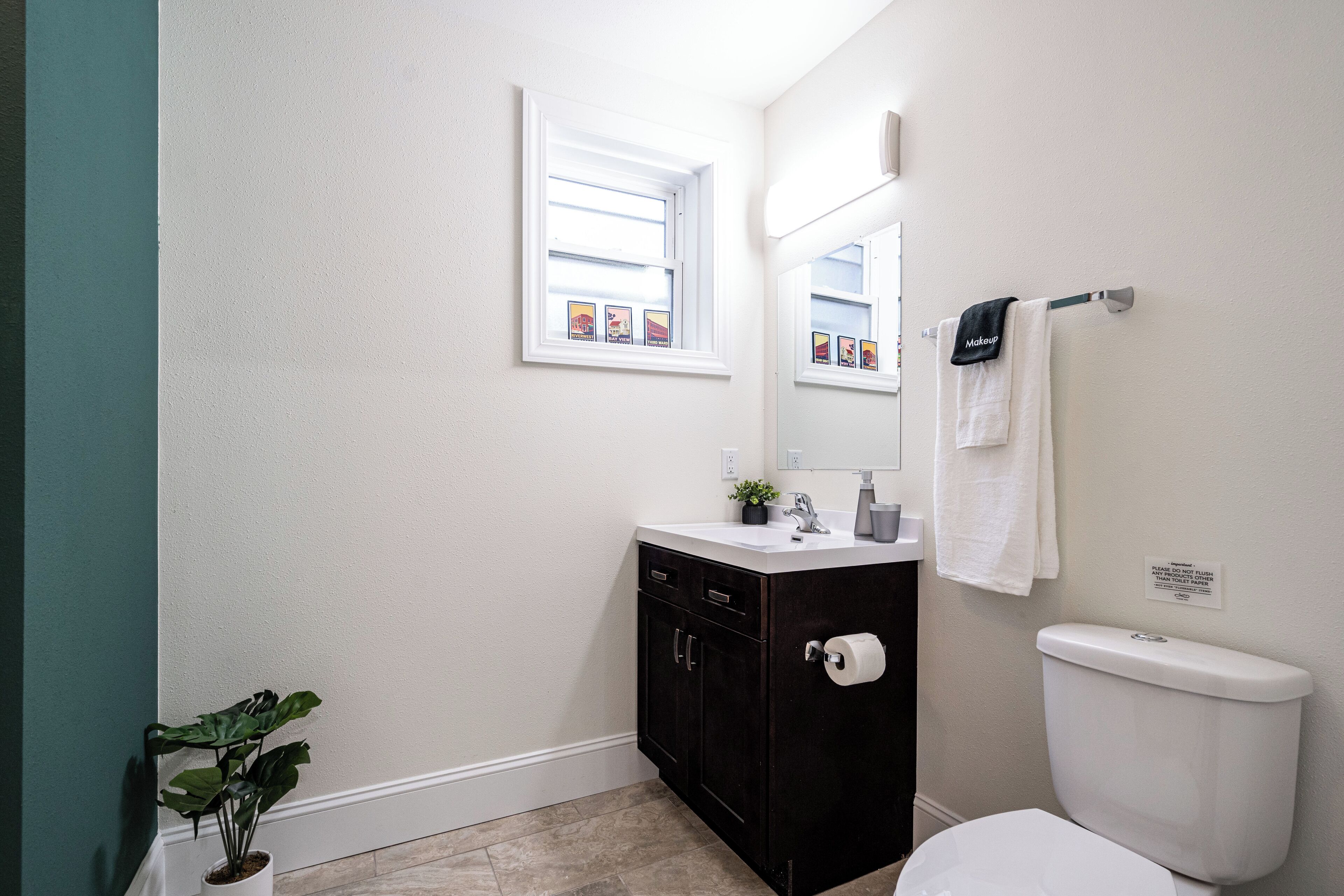 Un baño con lavamanos blanco, mueble oscuro, inodoro y ventana con una obra de arte.