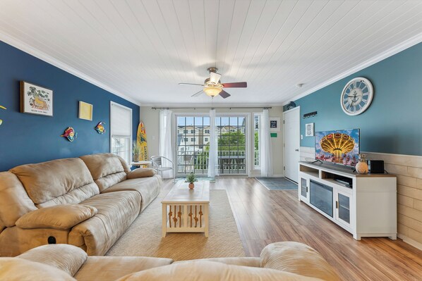 Un salon avec un canapé beige, une table basse, un ventilateur de plafond et une vue sur l’extérieur, visible à travers une porte vitrée.