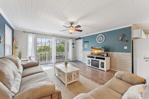 Un salon avec un ventilateur de plafond, un canapé, une table basse, un téléviseur posé sur un meuble et une vue sur l’extérieur par une porte vitrée.