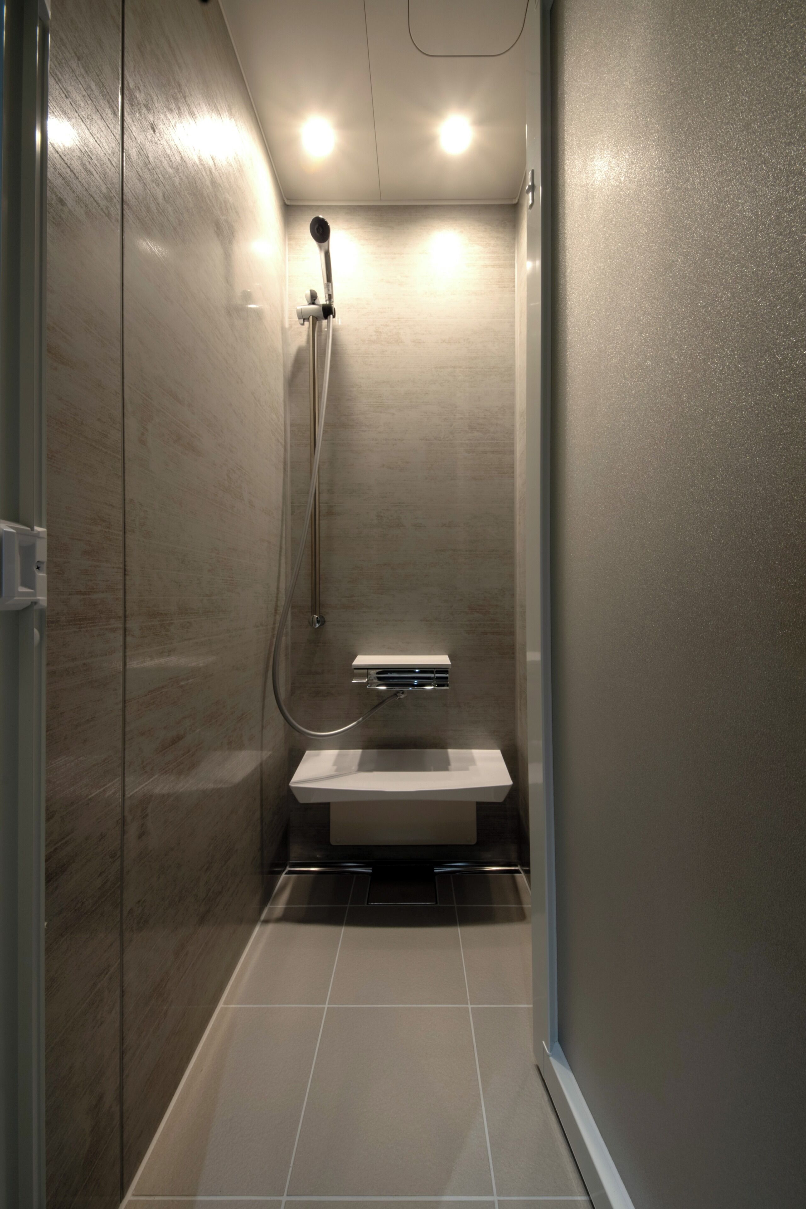 A narrow, enclosed shower area with a built-in toilet, tiled floor, and a showerhead mounted on the wall.