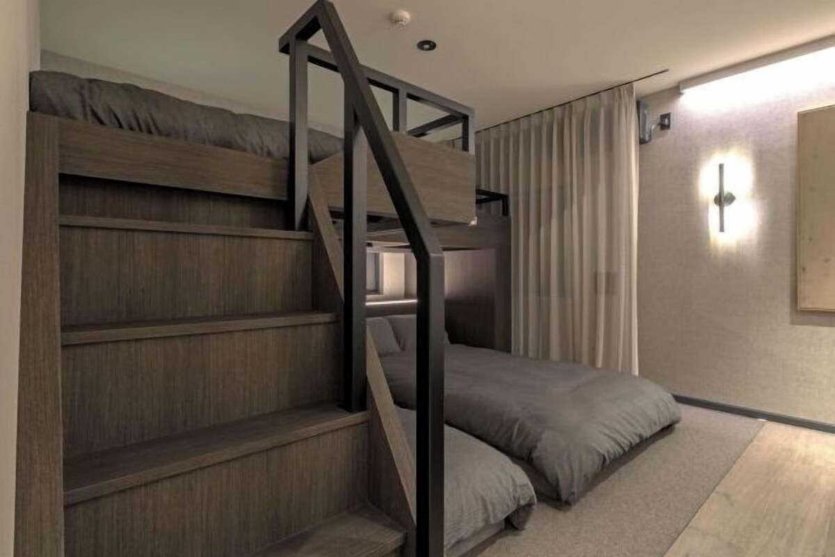 A bunk bed with a wooden ladder, a single bed with a pillow, a light switch on the wall, and a window with curtains.
