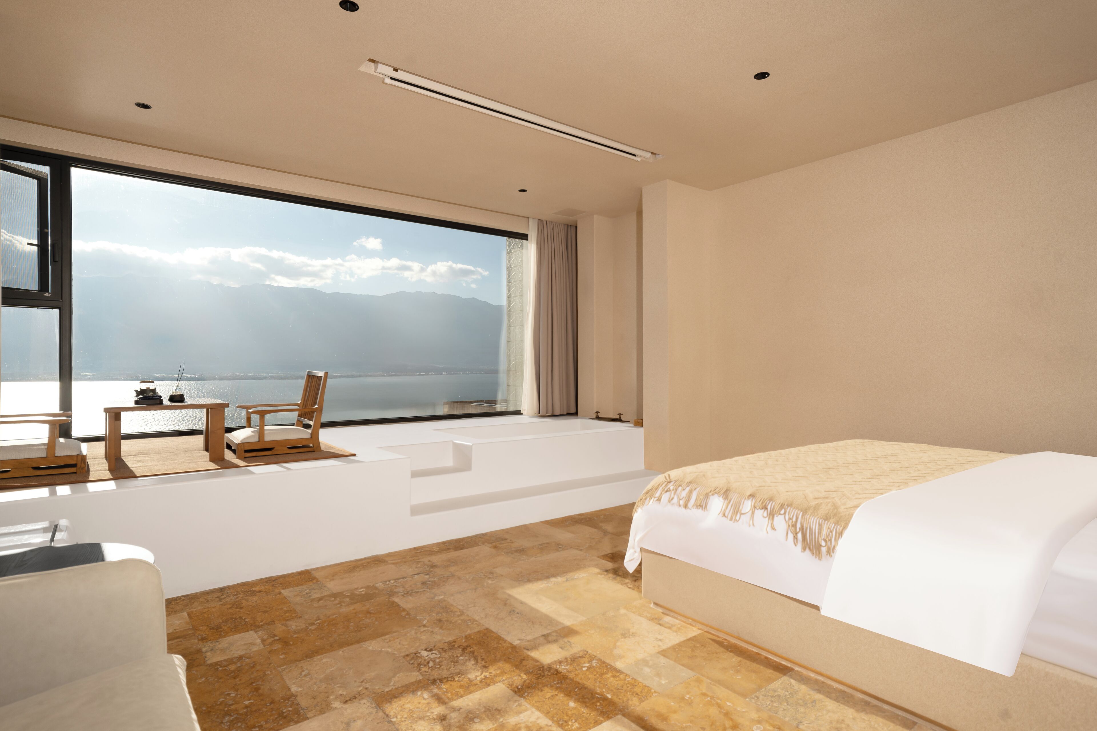 Una habitación de hotel moderna con un gran ventanal, una cama, un área de descanso con una mesa y sillas, y vista al mar.