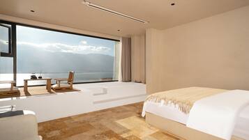 Una habitación de hotel moderna con un gran ventanal, una cama, un área de descanso con una mesa y sillas, y vista al mar.