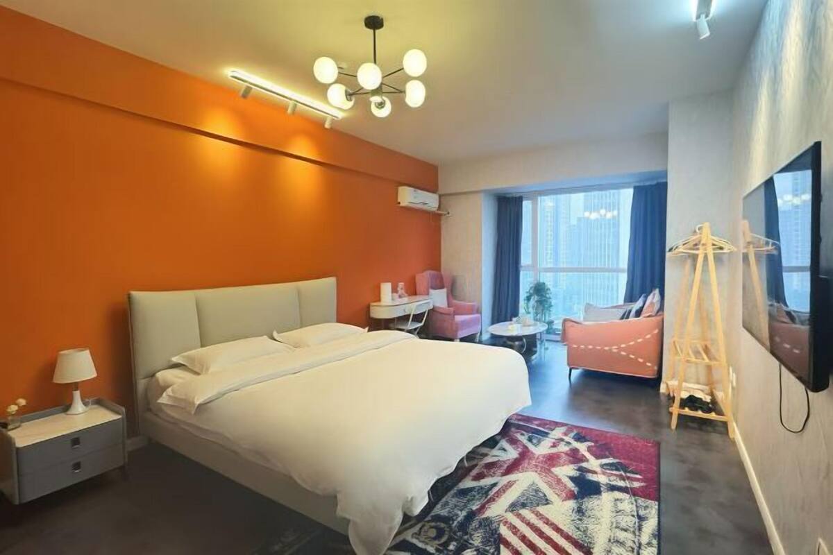 Ein modernes Hotelzimmer mit einem großen Bett, einem Nachttisch mit Lampe, einem kleinen Tisch mit Stuhl, einem farbenfrohen Teppich und einem großen Fenster mit Blick auf die Stadt.