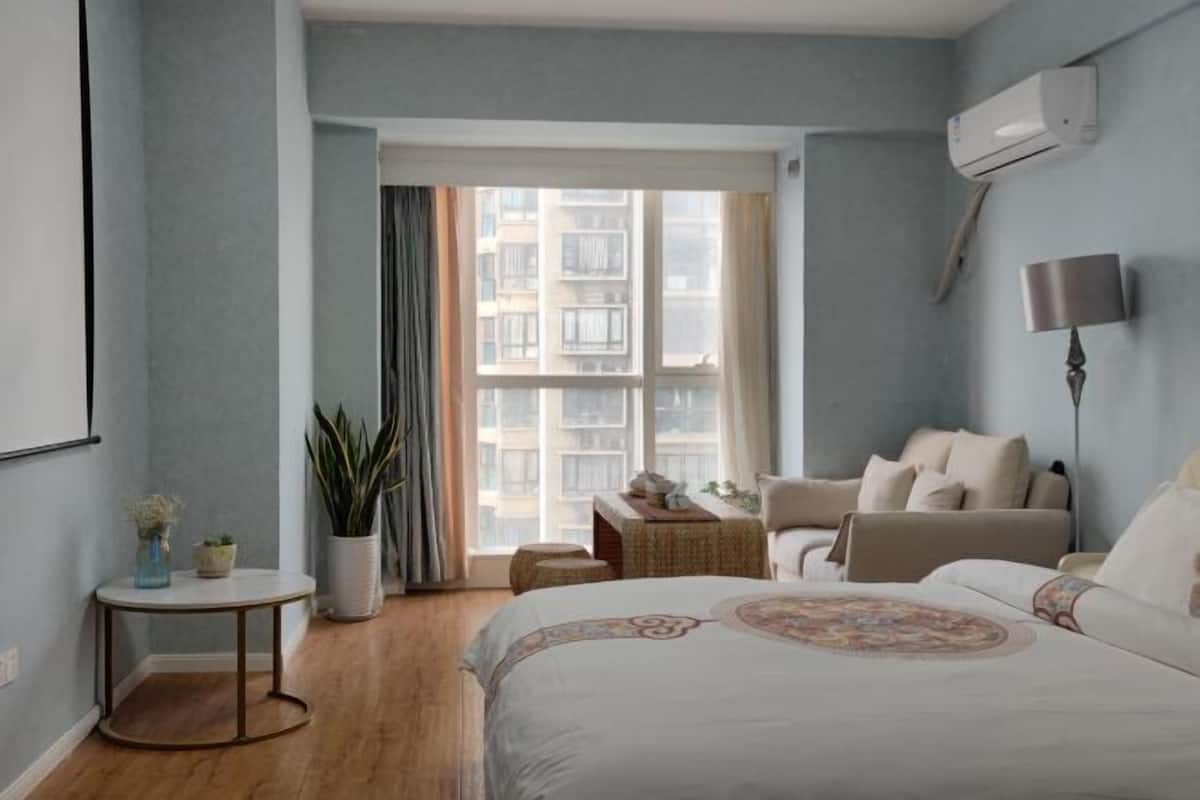 Ein Schlafzimmer mit einem Bett, einem Sessel, einem kleinen Tisch, einer Topfpflanze und einem Fenster mit Blick auf Gebäude.