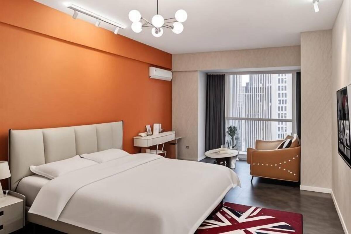 Ein Hotelzimmer mit einem großen Bett, einem Nachttisch, einem kleinen runden Tisch, einem Sessel und Blick auf die Stadt durch große Fenster.