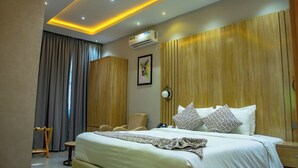 Een hotelkamer met een groot bed, een houten hoofdbord, een bureau met stoel, een kledingkast en een airconditioner.