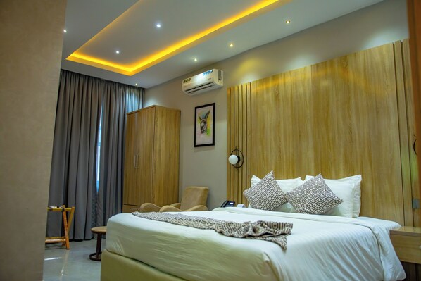 A hotel room with a large bed, a wooden headboard, a desk with a chair, a wardrobe, and an air conditioning unit. - Ojaja Suites Akure (Akure)