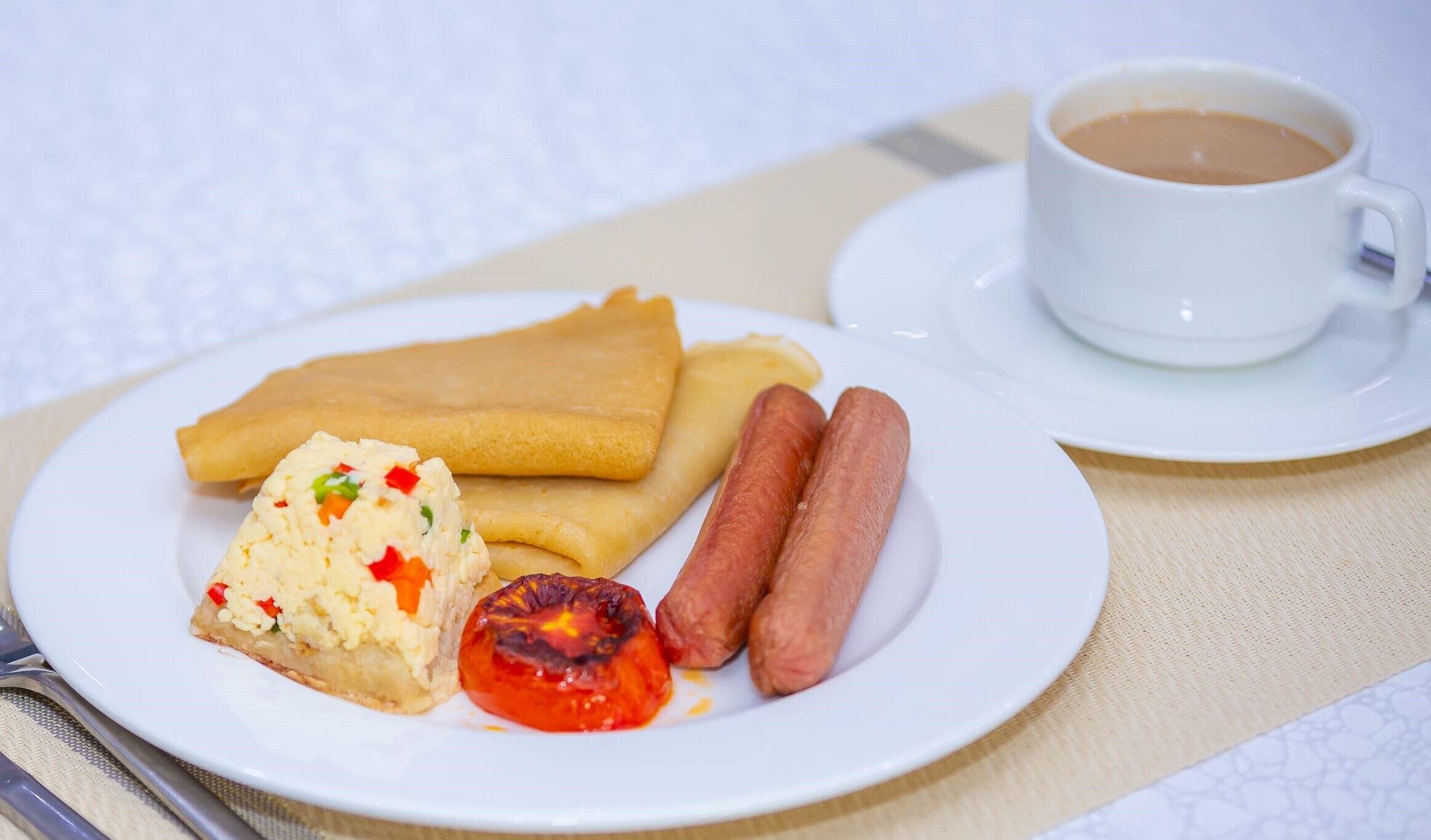 Une assiette contenant des crêpes, des saucisses, une tomate grillée et une tasse de café, posée sur une table.
