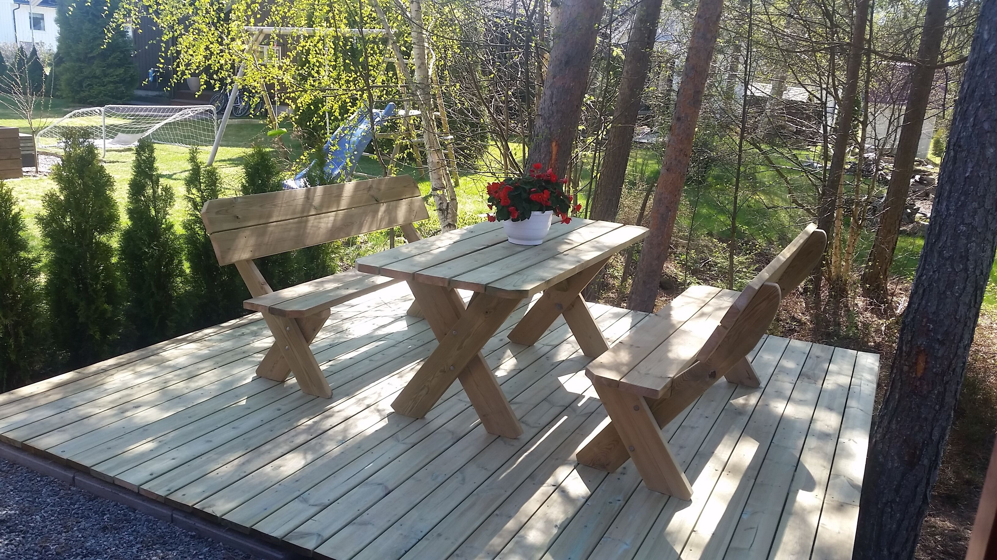 Una terraza de madera con una mesa de picnic y bancos, rodeada de árboles y vistas a un parque infantil.