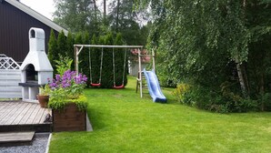 Ein Garten mit einer Holzterrasse, einem Grill, einer Schaukel und einer Rutsche.