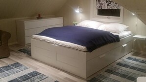A bedroom with a sloped ceiling, a bed with white and blue bedding, a bedside table with a lamp, and a window with a floral pattern. - Cozy Loft Close to the Nature (Ovre Eiker)