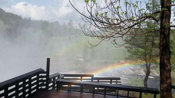 Eine Holzterrasse mit einer Bank und einer Aussichtsplattform, die auf einen bewaldeten Bereich mit einem sichtbaren Regenbogen blickt.