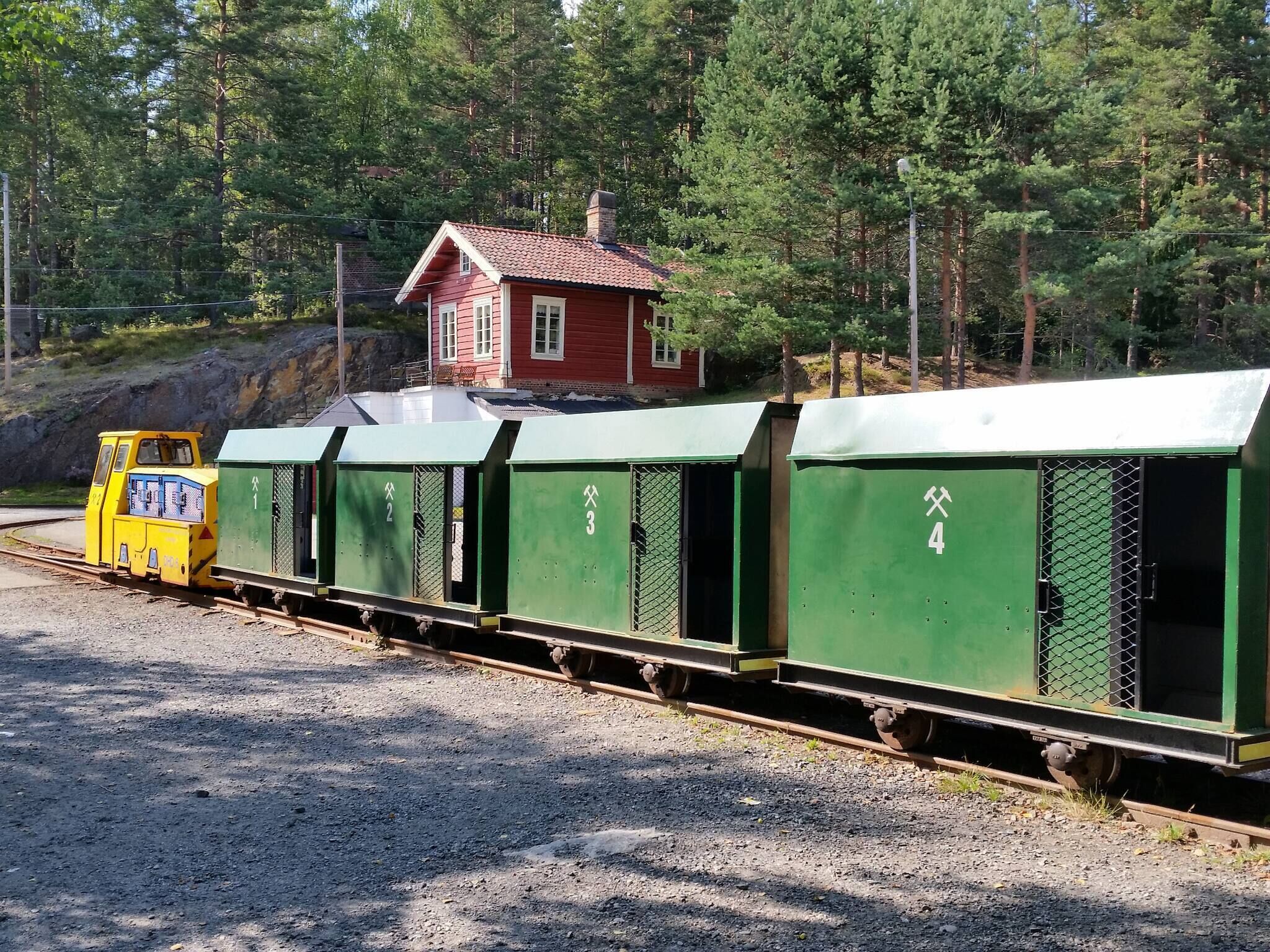 Un train composé de wagons de marchandises verts, numérotés de 1 à 4, d’une locomotive jaune et d’un bâtiment rouge en arrière-plan.