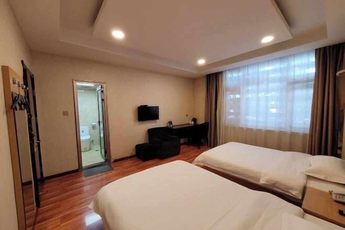 Habitación de hotel con dos camas, un escritorio con una silla, un televisor y un baño visible a través de una puerta de cristal.