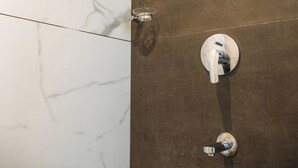 Un baño con piso de mármol y pared marrón. Hay tres elementos cromados: un cabezal de ducha, un grifo y un secador de manos.