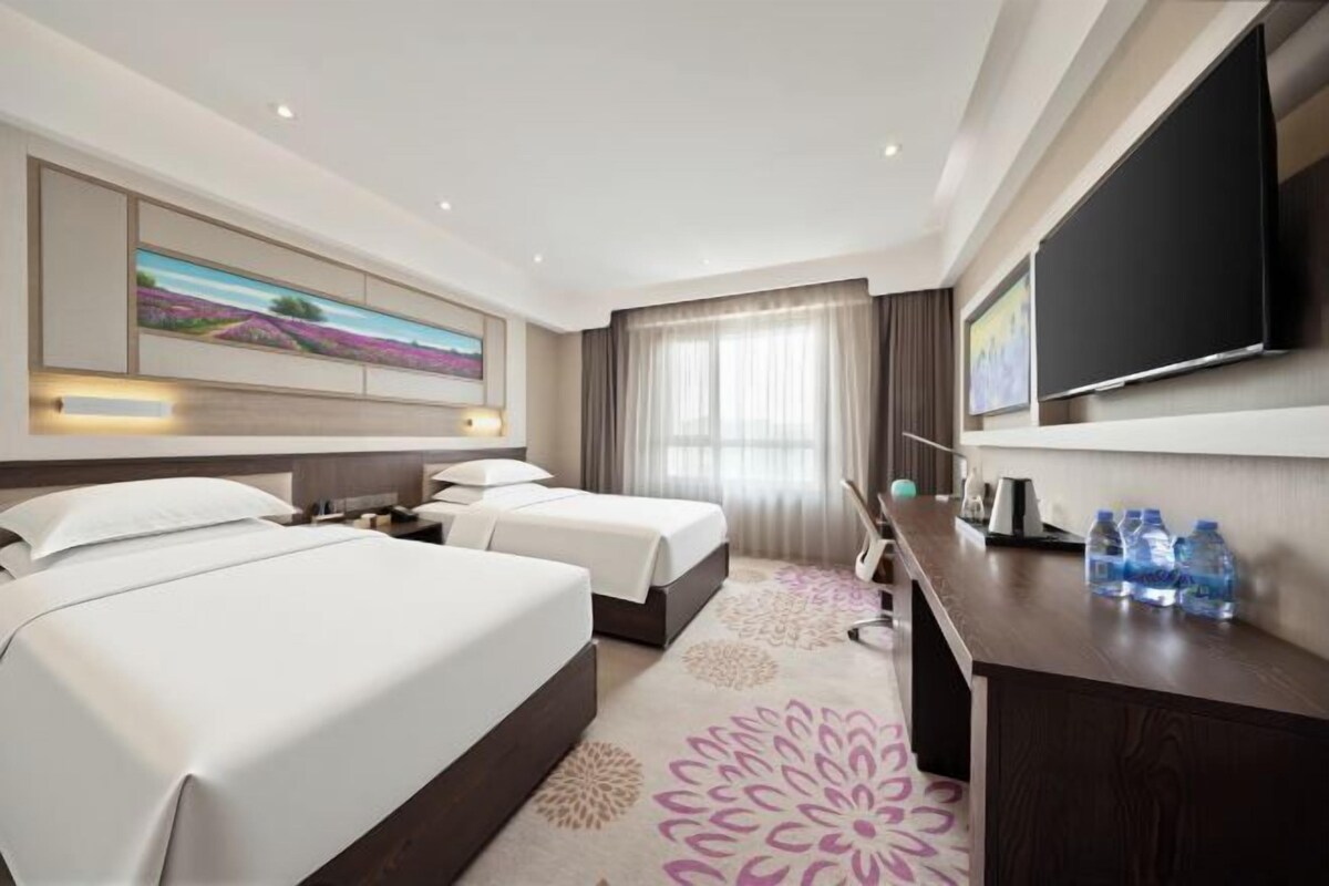 A hotel room with two beds, a desk, a flat-screen TV, and a large window with curtains.