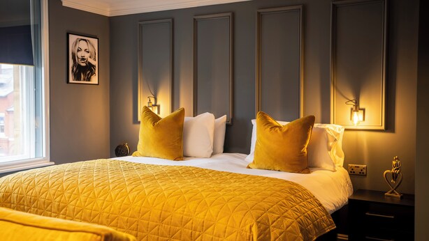 Una cama bien hecha con colcha amarilla, almohadas blancas y una mesita de noche con lámpara.