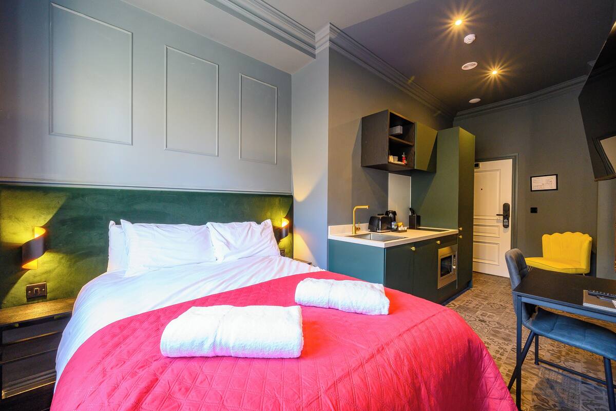 A hotel room with a large bed, a desk, a chair, a small kitchenette, and a bathroom.