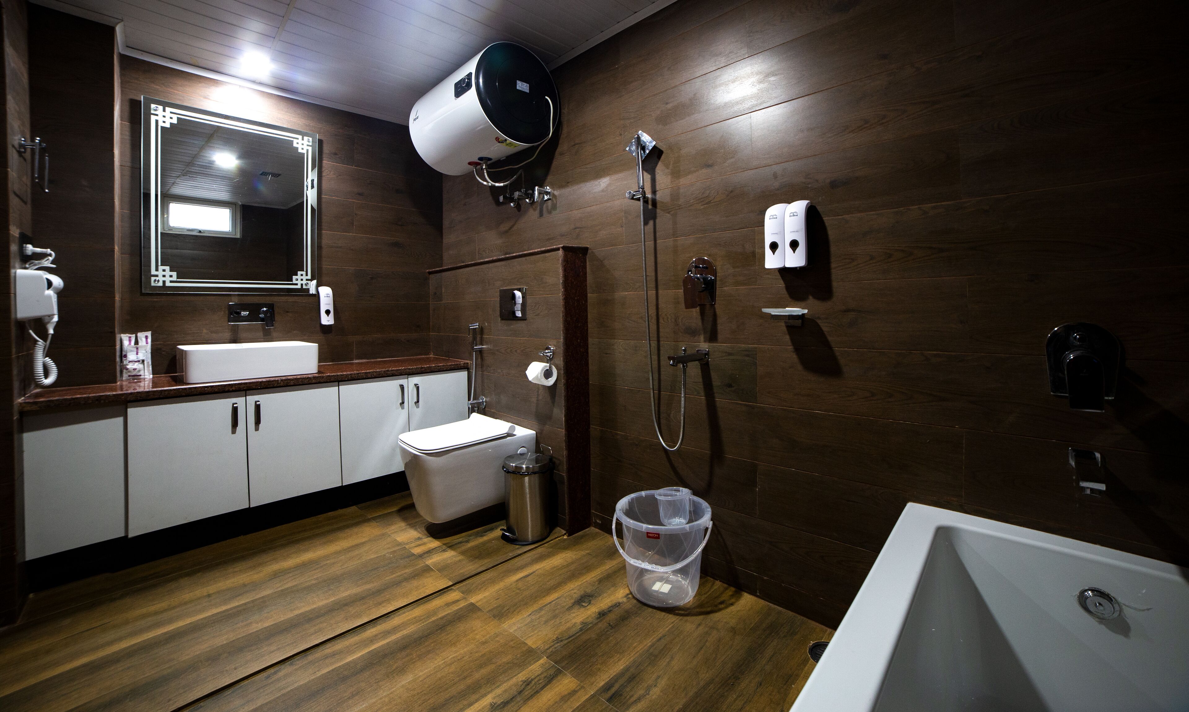 Un baño moderno con suelo de madera, un lavamanos blanco, inodoro, ducha y un espejo grande.