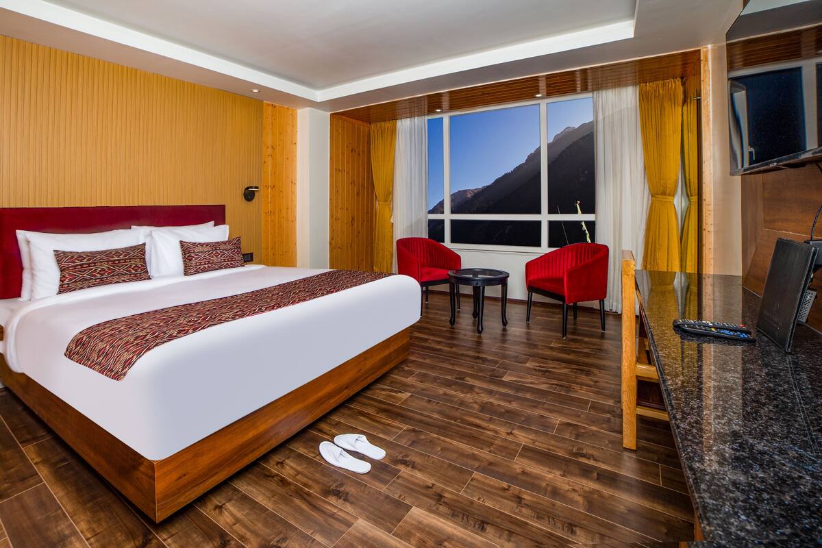A hotel room with a large bed, a small table, two red chairs, a TV, and a window with a mountain view.