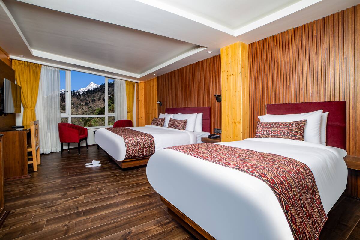 A hotel room with two beds, wooden walls, a large window with a mountain view, and a wooden desk with a chair.