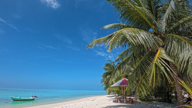 A beach with white sand, a clear blue sky, and a green boat in the water. - Ever Blue Guest House (Mathiveri)