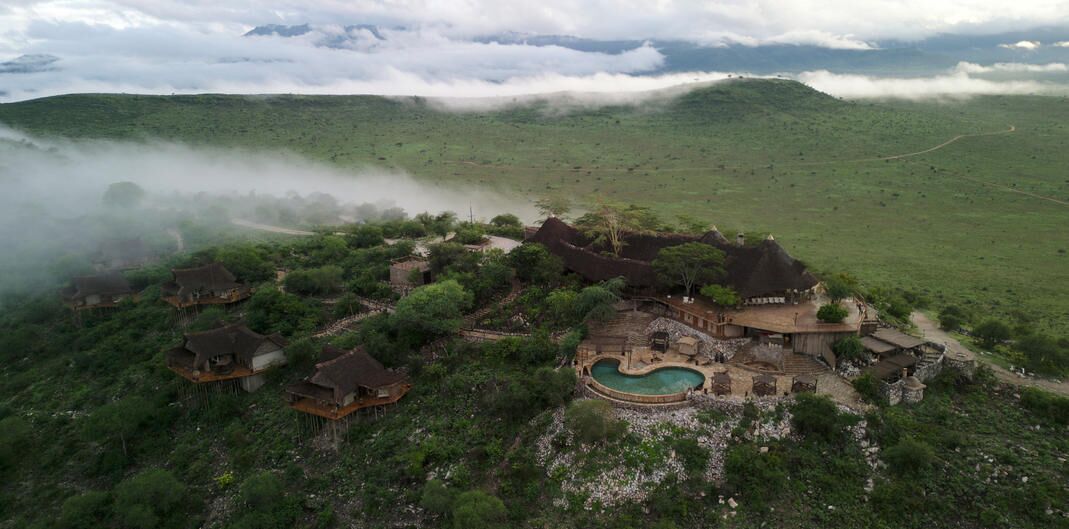 Un pueblo rural con casas tradicionales de techo de paja, un edificio central de piedra y una piscina rodeada de vegetación.