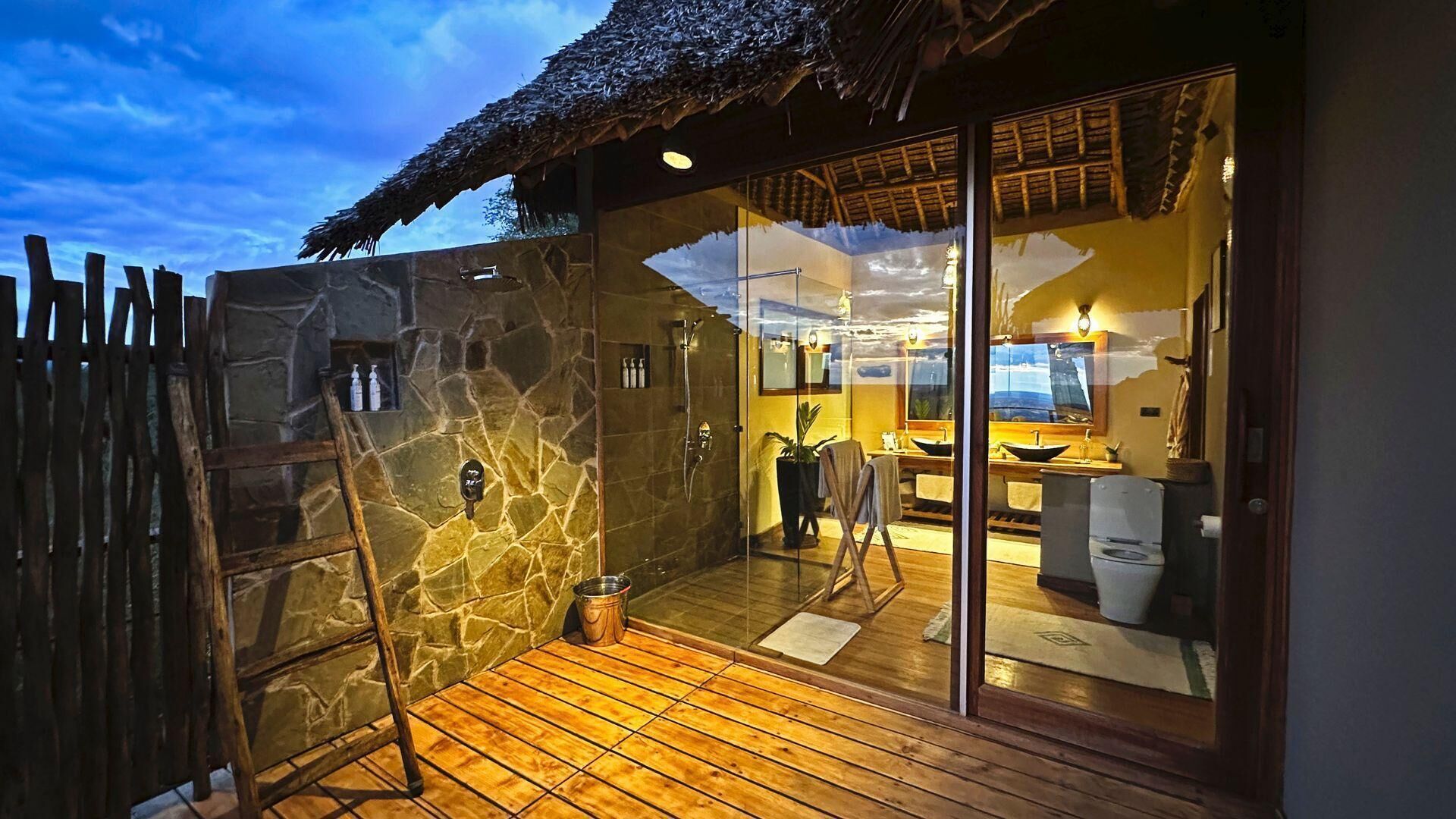 Une salle de bain aux murs en pierre, dotée d’une douche en verre, d’une terrasse en bois et d’un toit de chaume.