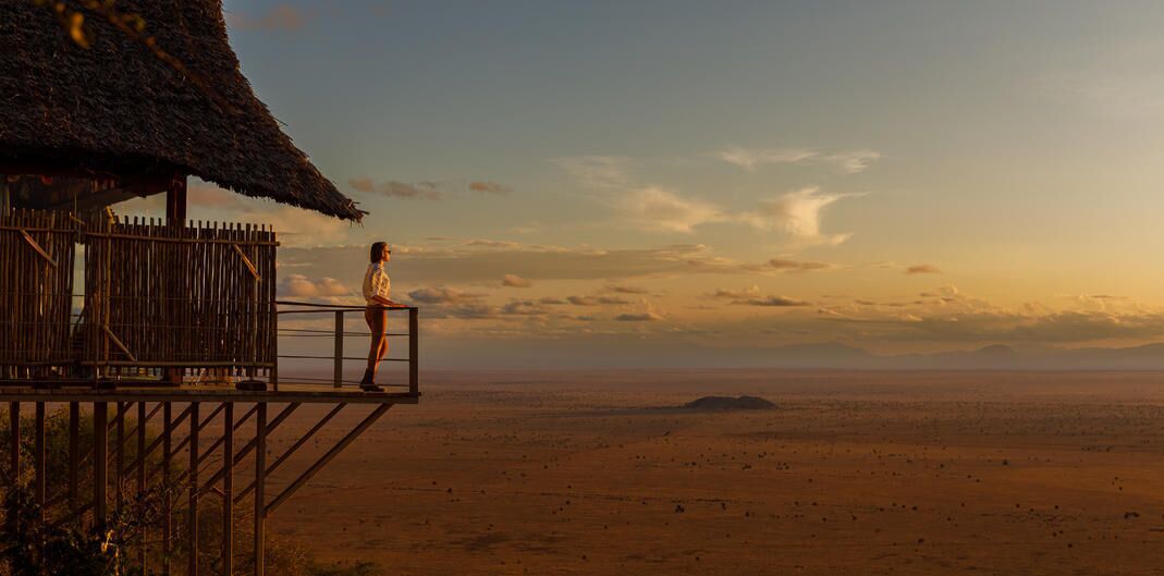 A person standing on a wooden deck overlooking a vast desert landscape with a thatched-roof hut.