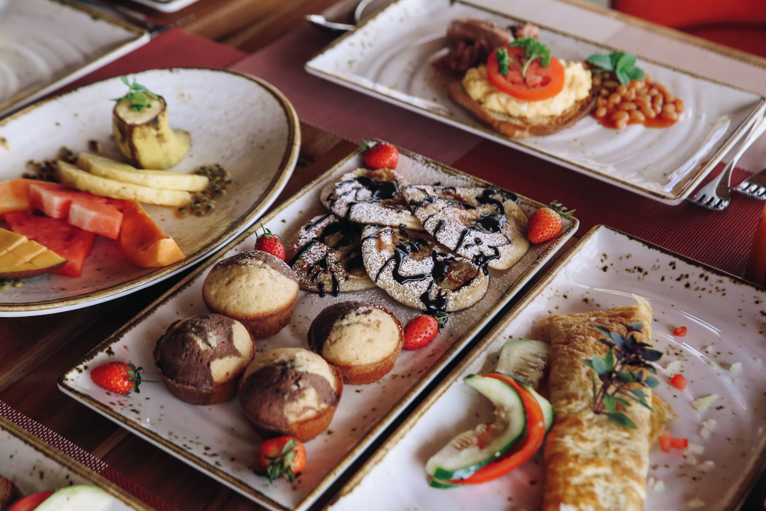A variety of plated dishes including pancakes, muffins, and a rolled omelette, arranged on a table.
