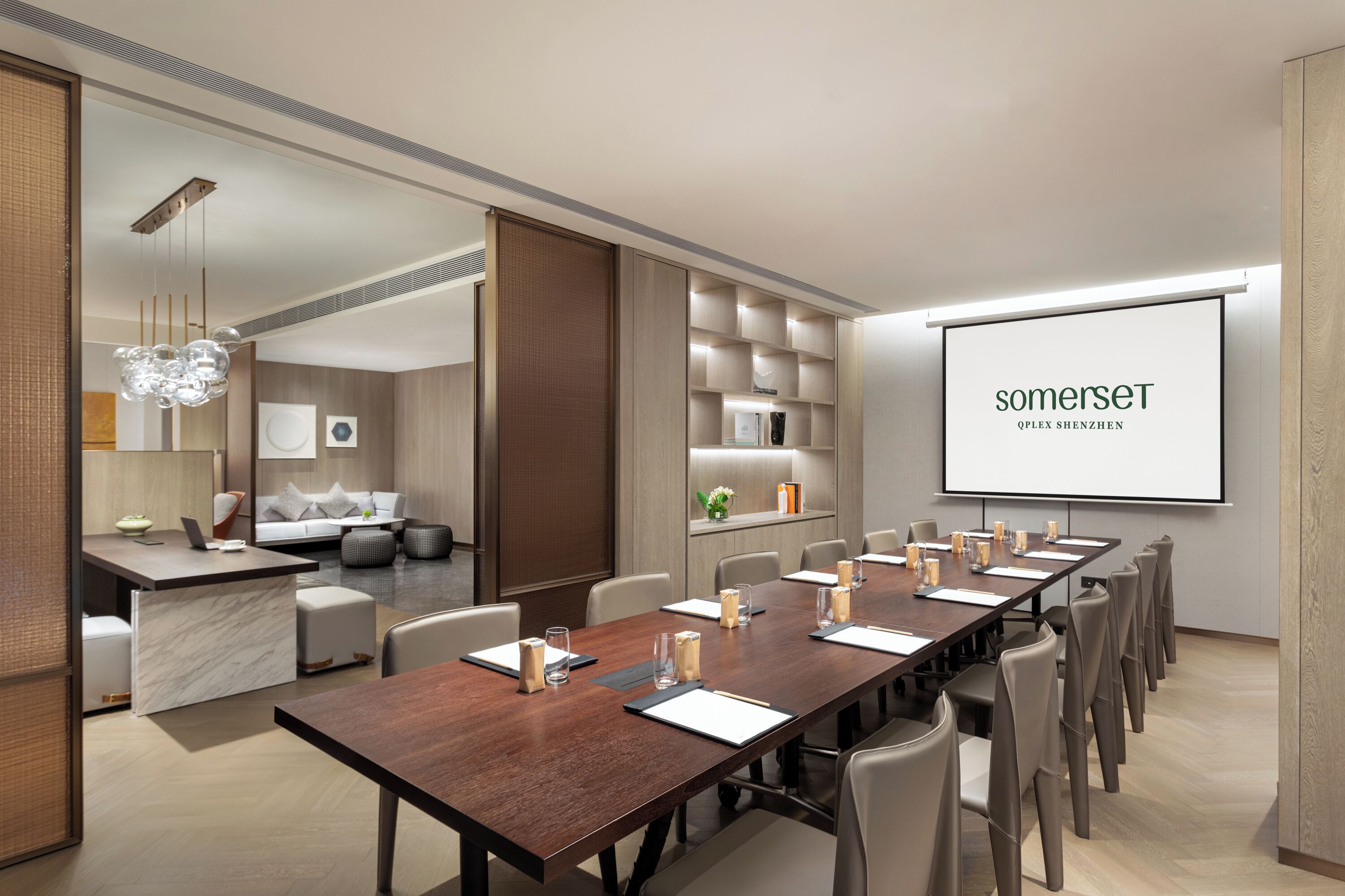A modern conference room with a long table, chairs, a projector screen, and a seating area with a sofa and coffee table.