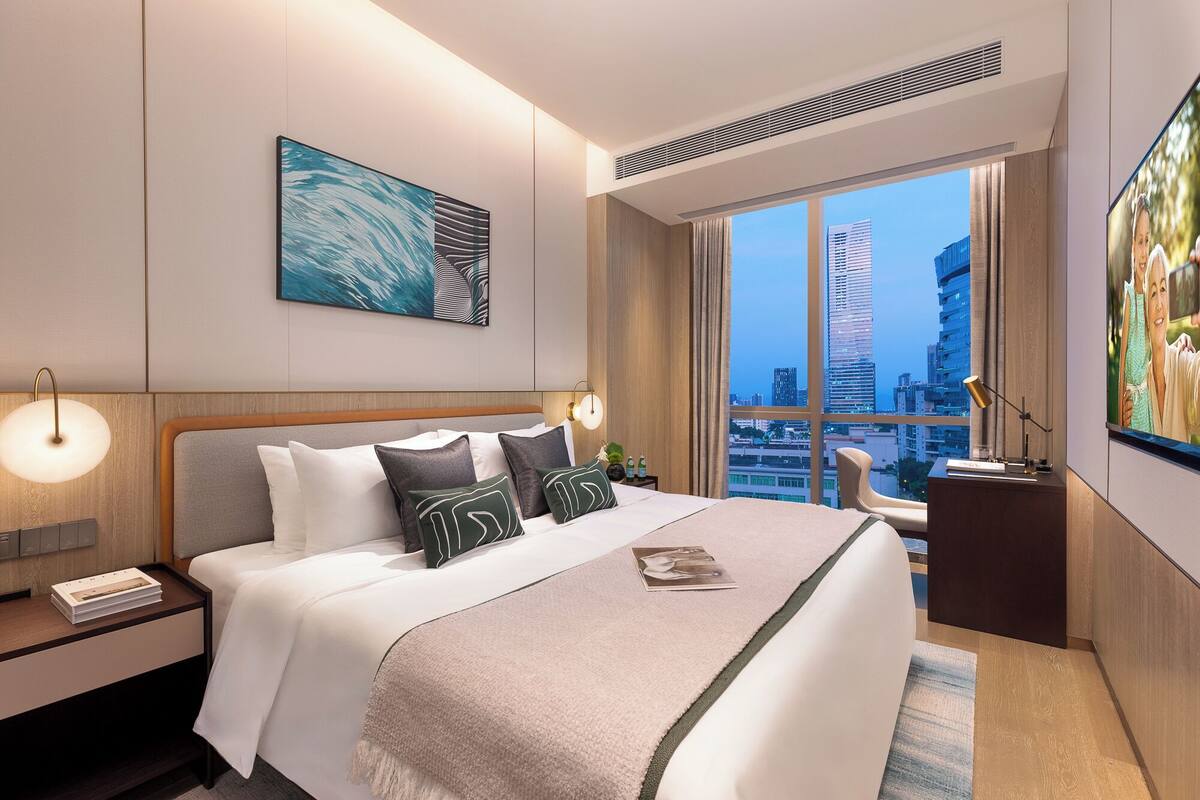 A modern hotel room with a large bed, a city view, a painting on the wall, and a desk with a lamp.