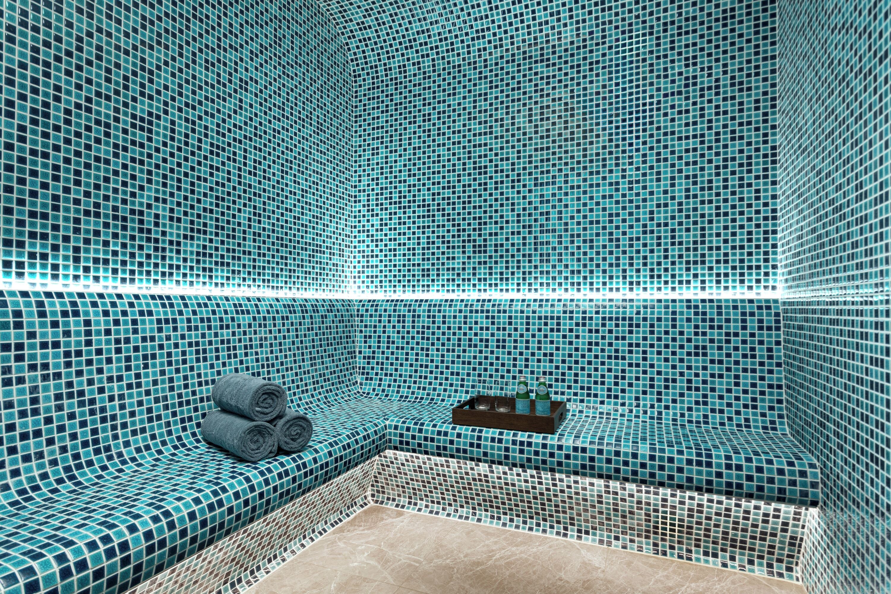 A bathroom with a tiled mosaic wall, a bench with towels, and a tray with bottles.
