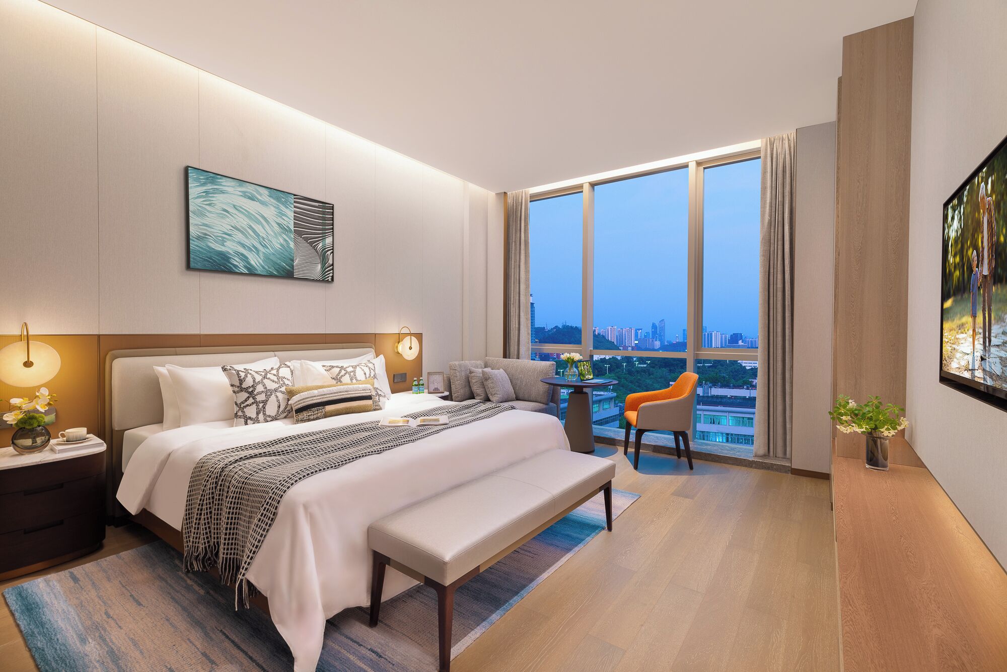 A modern hotel room with a large bed, a desk, a chair, and a view of the cityscape through large windows.