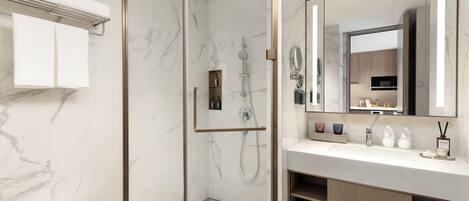 Un baño moderno con ducha de vidrio, lavamanos blanco y amplio espejo, y paredes de mármol.