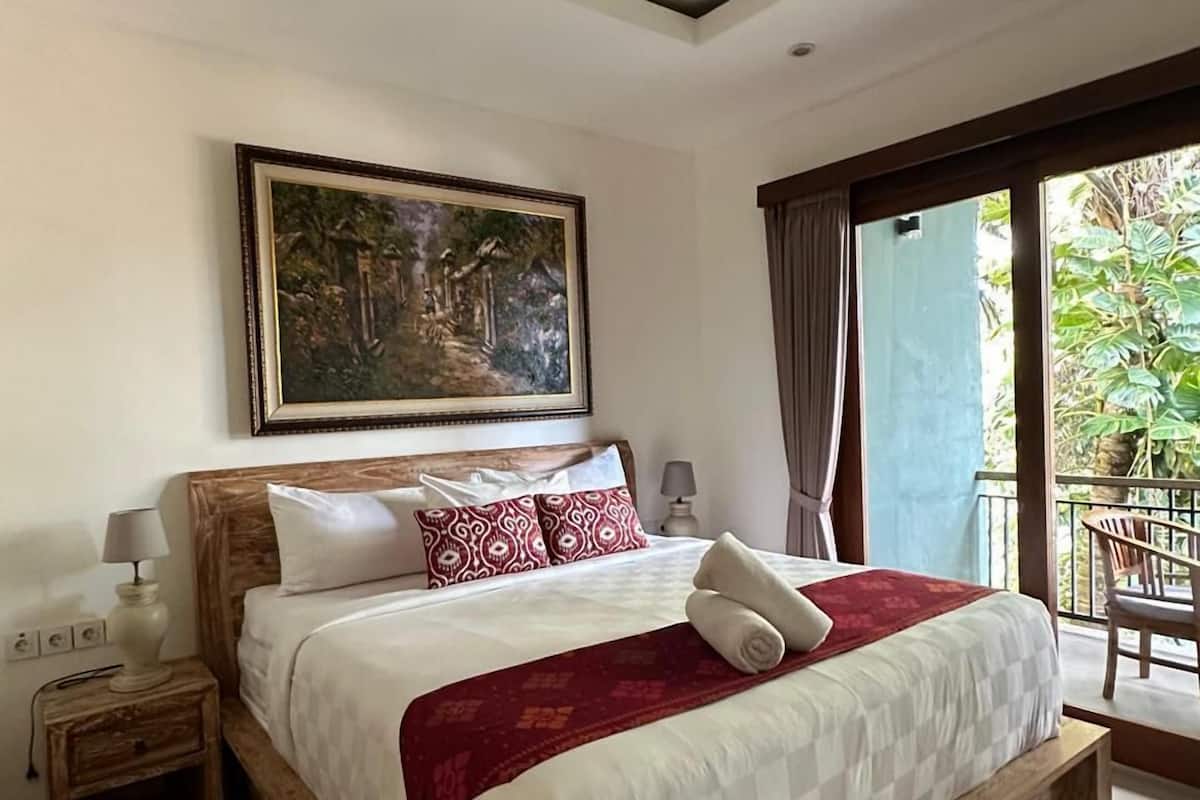 A modern bedroom with a large bed, a wooden headboard, a painting above the bed, and a balcony with a view of greenery.