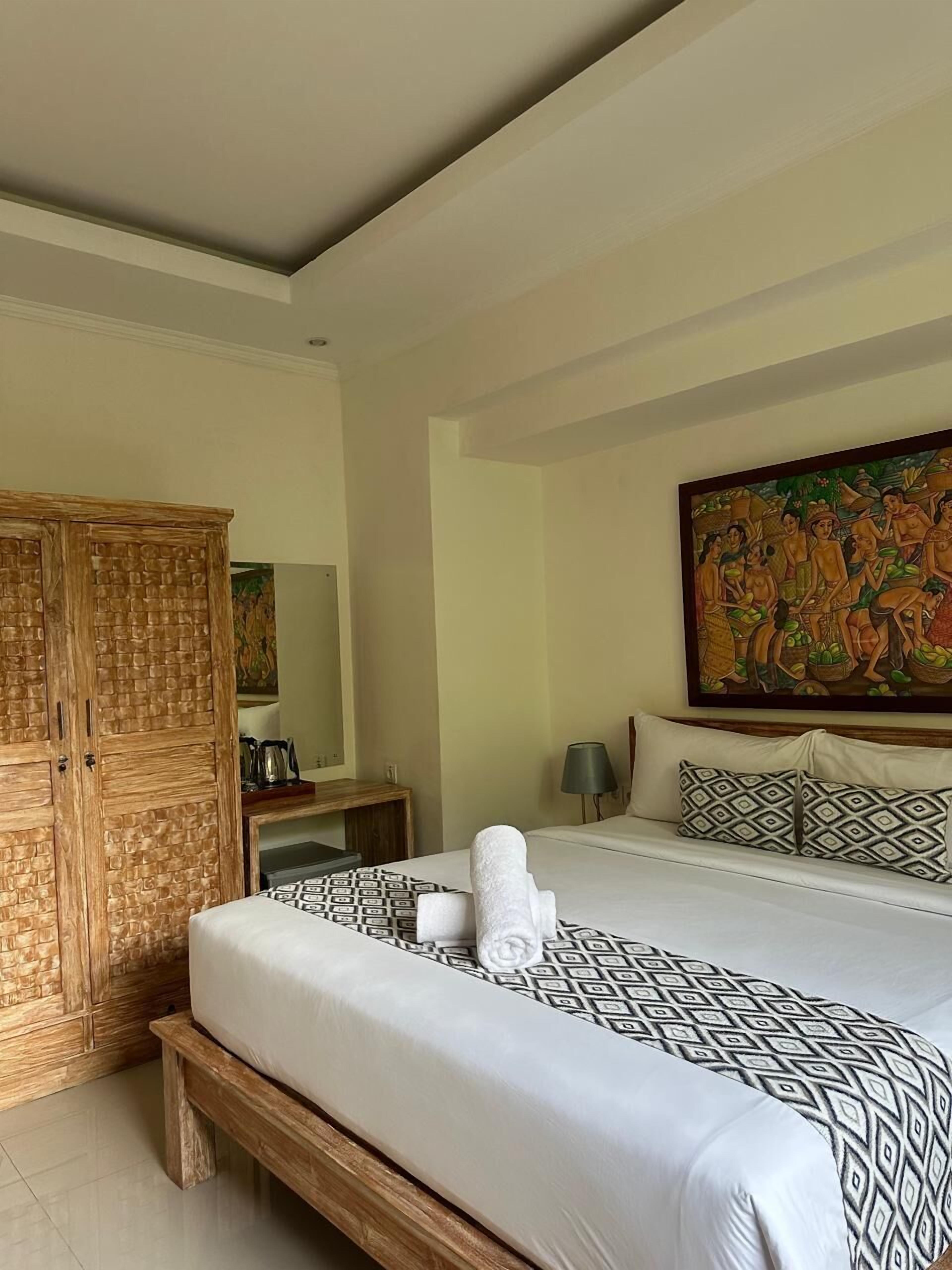 A hotel room with a large bed, a wooden wardrobe, a painting on the wall, and a bedside table with a lamp.