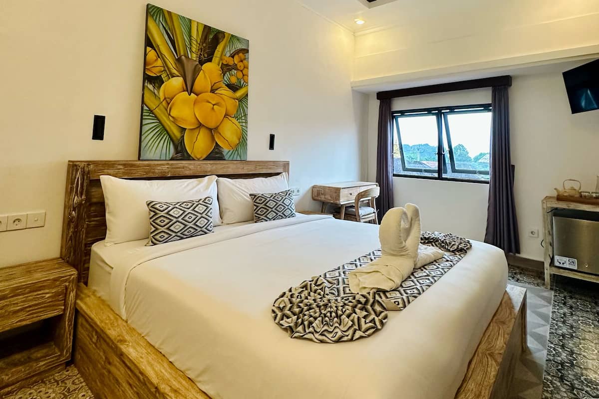 A hotel room with a large bed, a wooden headboard, a painting of yellow coconuts, a window with curtains, and a small table with a TV.