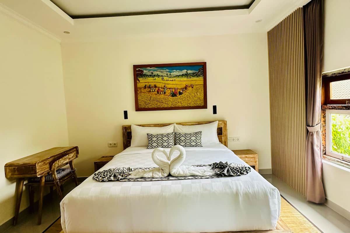 A bedroom with a large bed, a wooden headboard, a painting above the bed, a bedside table, a bench, and a window with curtains.
