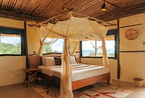 Un dormitorio con una cama de madera, una mesita de noche, una cesta tejida y vistas al exterior a través de amplias ventanas.