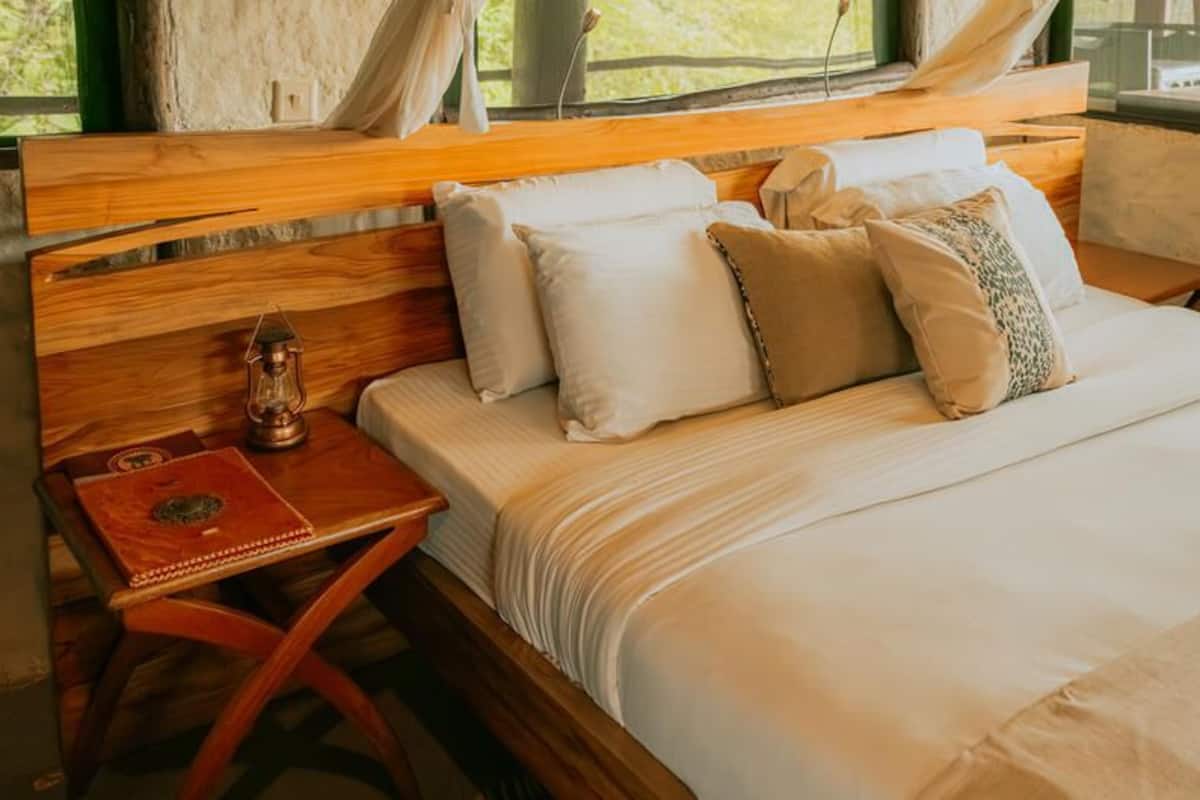 Ein Bett mit Holz-Kopfstück, ein kleiner Holztisch mit einer Laterne und ein Fenster mit Vorhängen.